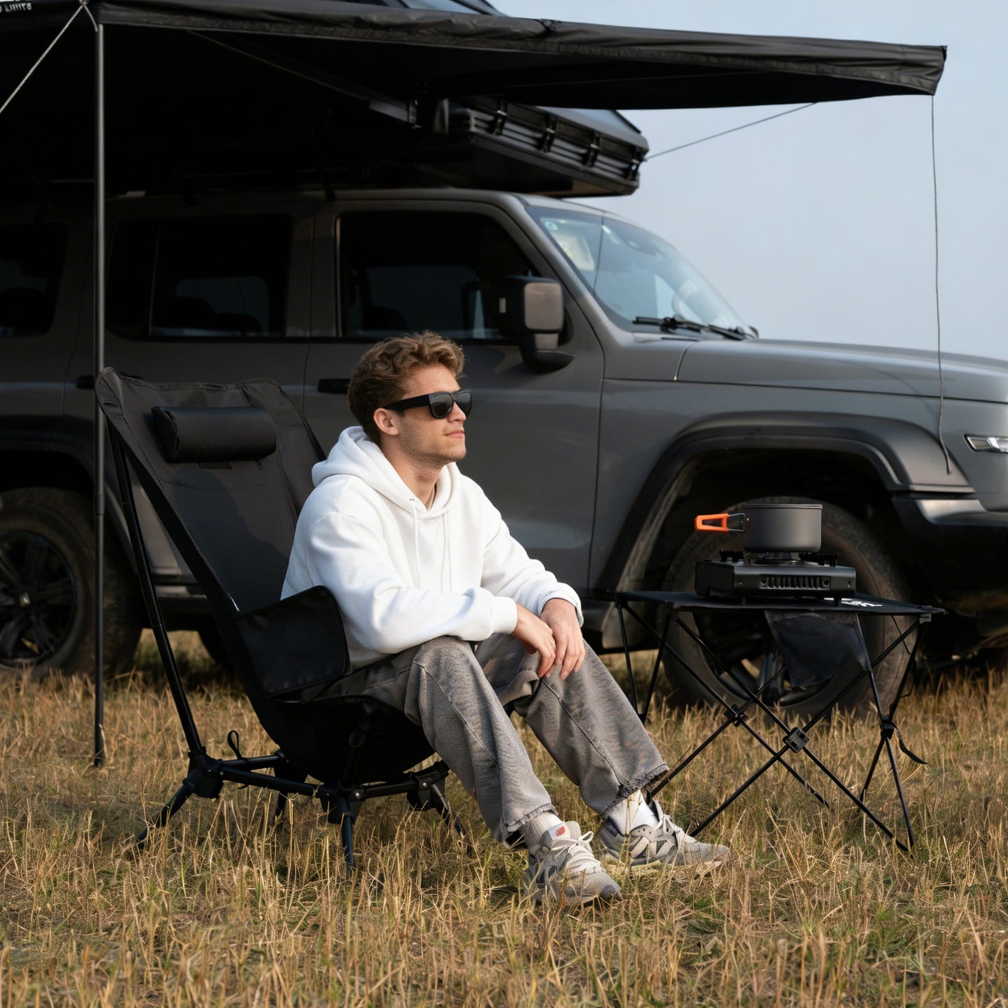 Person sitting on a camping chair under an awning beside a vehicle and portable table in an outdoor campsite.