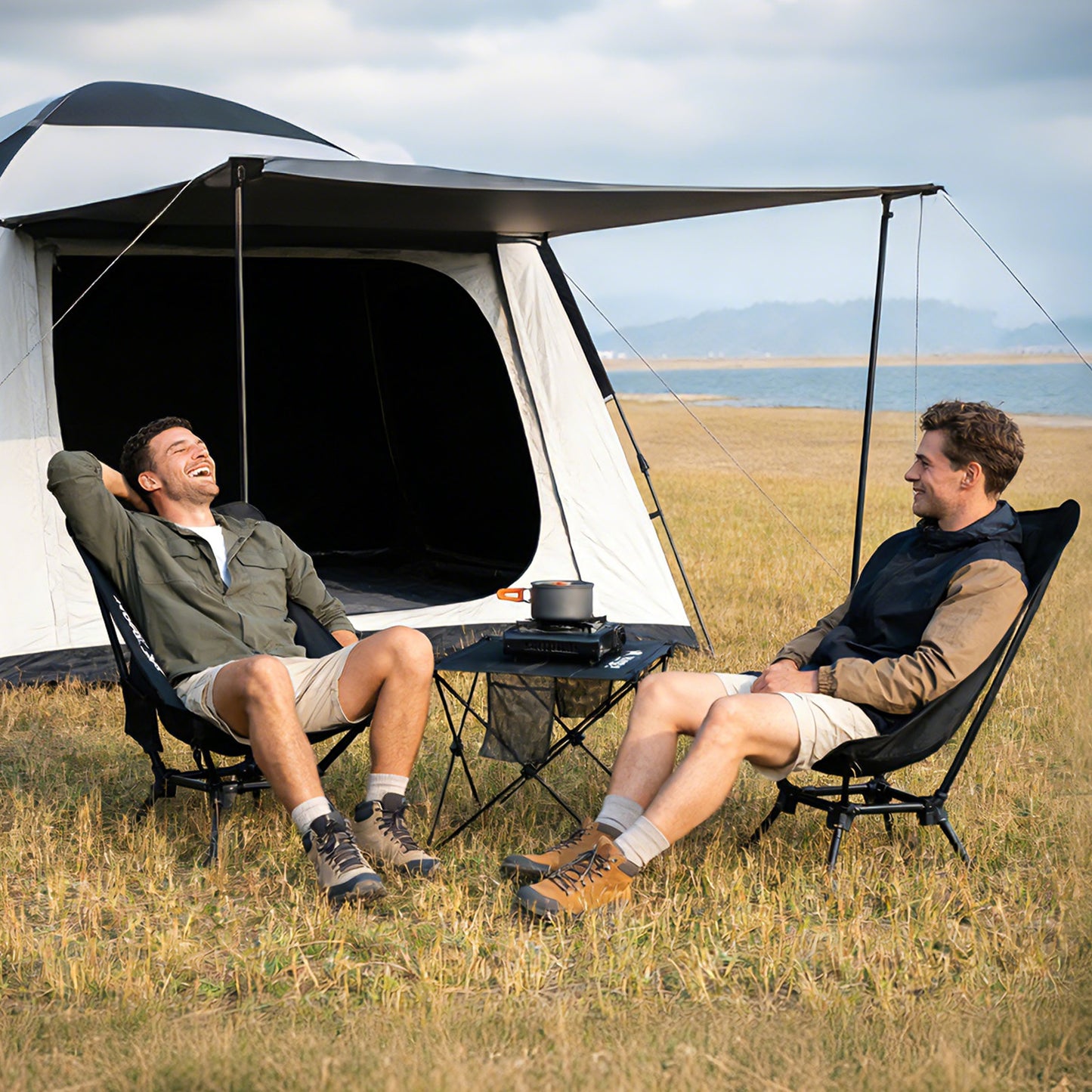 Two people relaxing on camping chairs beside a tent and folding table, enjoying an outdoor campsite setup.