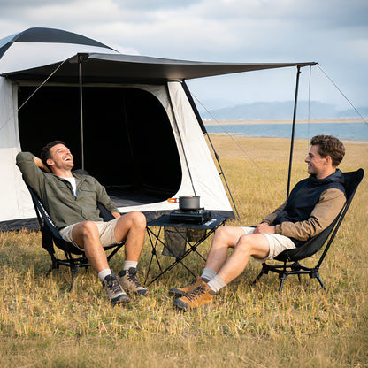Two people relaxing on camping chairs beside a tent and folding table, enjoying an outdoor campsite setup.