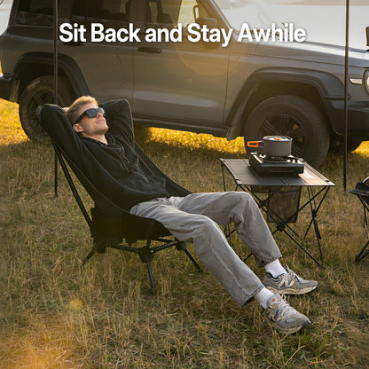 Person relaxing on a reclining camping chair beside a portable table and vehicle at an outdoor campsite.